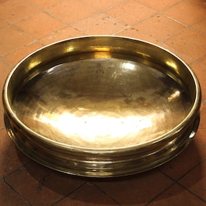 May include: A large, round, brass bowl with a wide, shallow basin and a series of concentric rings around the sides. The bowl has a polished, reflective surface, and sits on a brown tiled surface.