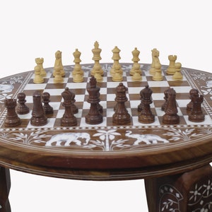 May include: A round wooden chess table with intricate carvings of elephants and floral designs. The table has three legs and a chessboard with a set of chess pieces.