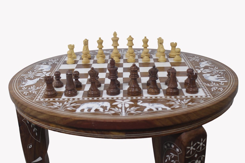 May include: A round wooden chess table with intricate carvings of elephants and floral designs. The table has three legs and a chessboard with a set of chess pieces.