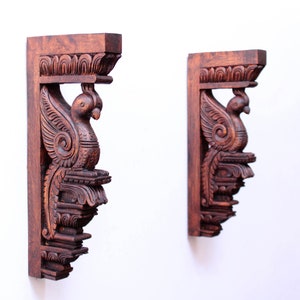 May include: Two intricately carved wooden corbels, each featuring a bird with outstretched wings. The corbels are a rich brown color and have a traditional Indian design.