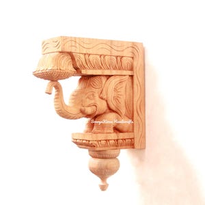 May include: A wooden wall bracket with an intricately carved elephant design. The bracket is made of light brown wood and has a traditional Indian style. The elephant is holding a decorative element in its trunk.