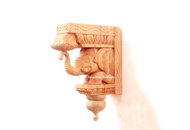 May include: A wooden wall bracket with an intricately carved elephant design. The bracket is made of light brown wood and has a traditional Indian style. The elephant is holding a decorative element in its trunk.
