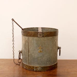 May include: A vintage metal bucket with a chain and a handle. The bucket is painted a light gray color and has a brass band around the top. The bucket has a cross design on the front.