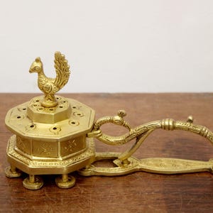 May include: A gold-colored incense burner with a peacock-shaped finial. The burner has an octagonal body with small holes and a handle. The piece is made of metal and sits on a wooden surface.