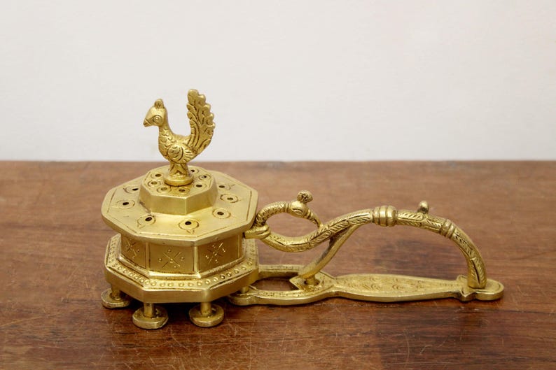 May include: A gold-colored incense burner with a peacock-shaped finial. The burner has an octagonal body with small holes and a handle. The piece is made of metal and sits on a wooden surface.