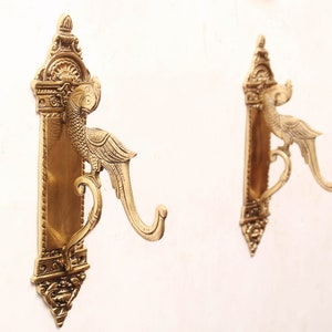 Op de afbeelding: Twee sierlijke messing wandhaken. Elke haak heeft een decoratief vogelontwerp met een gebogen haak om items op te hangen. De haken zijn gemonteerd op een rechthoekige achterplaat met ingewikkelde details. De haken zijn ongeveer 20 cm hoog.