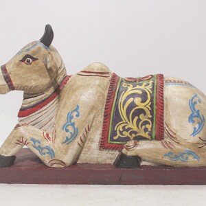 Puede incluir: Una escultura de madera pintada a mano de una vaca acostada. La vaca es blanca con detalles azules y rojos. La vaca lleva una silla de montar decorativa con diseños dorados y morados.