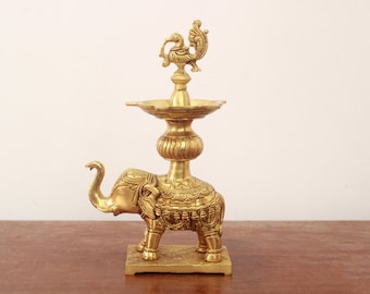 May include: A gold metal diya with an elephant base and a peacock finial. The diya has a round bowl for oil and wicks.