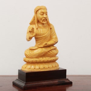 May include: A wooden statue of a seated figure with long hair and a beard, wearing a robe. The statue is painted in a light yellow color and is sitting on a lotus-shaped base. The statue is on a wooden base.