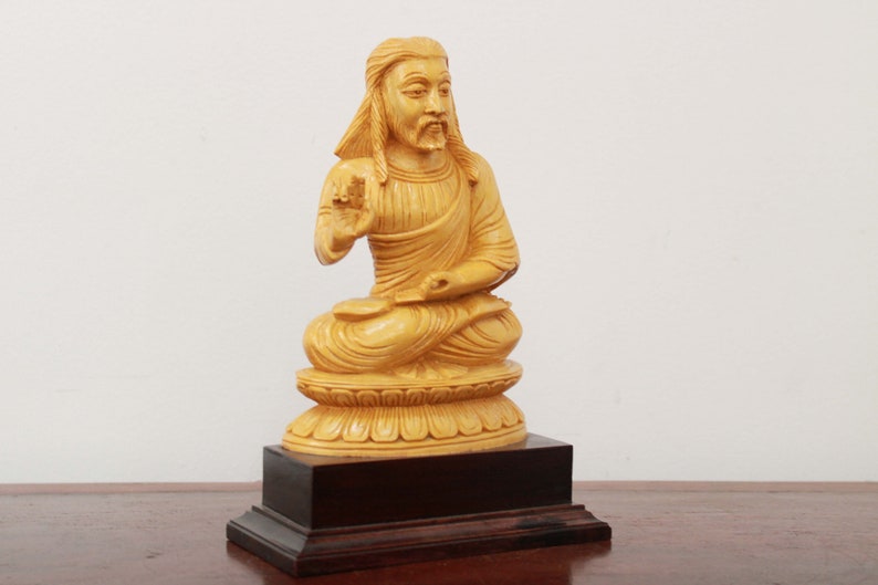 May include: A wooden statue of a seated figure with long hair and a beard, wearing a robe. The statue is painted in a light yellow color and is sitting on a lotus-shaped base. The statue is on a wooden base.