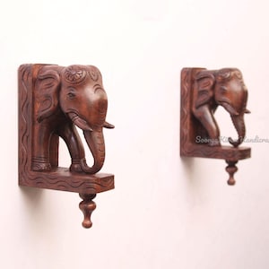 May include: Two dark brown wooden elephant wall shelves. Each shelf features a carved elephant with detailed features, including tusks and a decorative headpiece. The shelves have a small, decorative base with a turned finial.