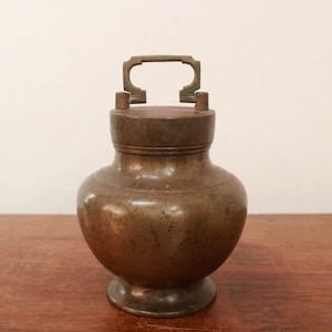 May include: A small, antique, bronze jar with a lid and a handle. The jar has a rounded body and a slightly flared base.