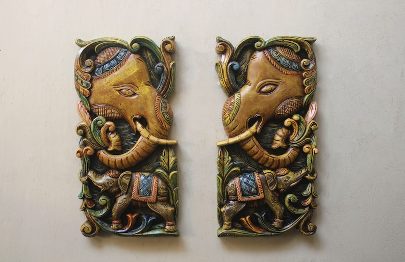 May include: Two intricately carved wooden wall hangings depicting elephants. The elephants are painted in shades of gold, brown, and green with intricate details. The carvings are surrounded by floral designs.