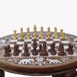May include: A round wooden chessboard table with intricate carvings of elephants and foliage. The chessboard is made of light and dark wood squares and features a set of wooden chess pieces in the middle of the board.