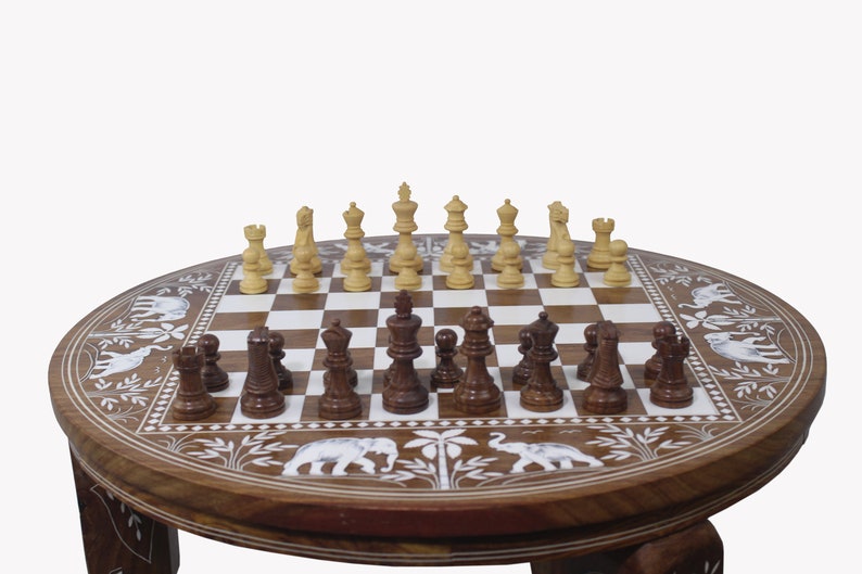 May include: A round wooden chessboard table with intricate carvings of elephants and foliage. The chessboard is made of light and dark wood squares and features a set of wooden chess pieces in the middle of the board.