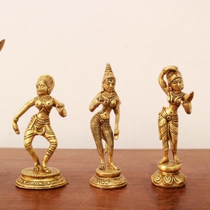 May include: Five gold-colored statues of female figures in various poses. Each statue is intricately detailed, depicting traditional attire and hairstyles. The statues are displayed on a wooden surface against a plain background, showcasing their elegant design.