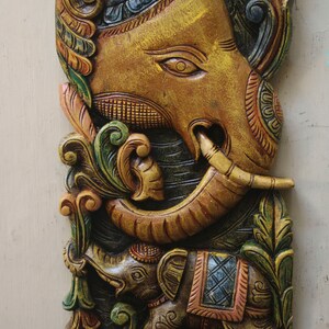 May include: A carved wooden wall hanging featuring a large, intricately detailed elephant with a smaller elephant at its feet. The elephants are painted in shades of brown, green, and blue, with intricate floral designs.