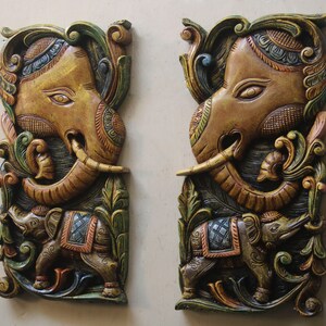 May include: Two intricately carved wooden wall decor pieces featuring elephant heads with ornate details. The elephants are painted in shades of gold, brown, green, and blue, with intricate floral patterns surrounding them.