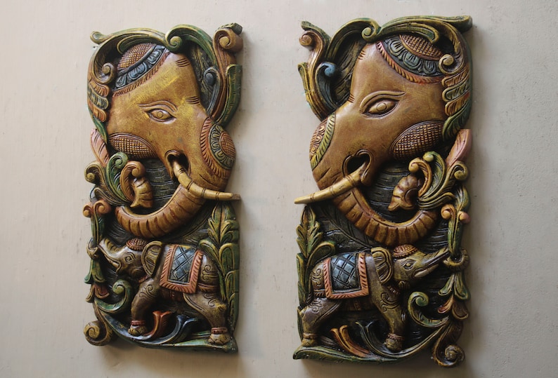 May include: Two intricately carved wooden wall decor pieces featuring elephant heads with ornate details. The elephants are painted in shades of gold, brown, green, and blue, with intricate floral patterns surrounding them.