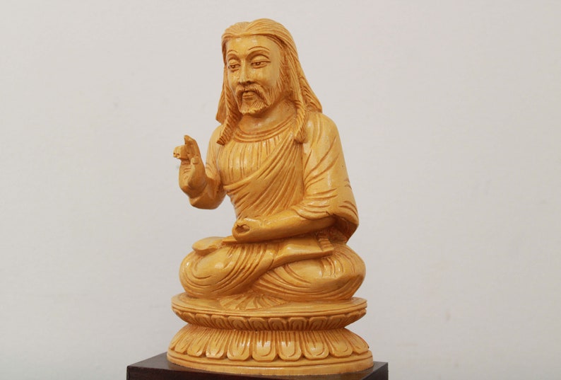 May include: A wooden statue of a person sitting in a meditative pose with their hands in their lap. The statue is carved from light brown wood and has a detailed face with a long beard.