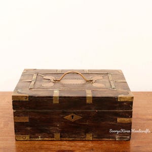 May include: A wooden box with brass accents and a copper inlay. The box has a hinged lid with a brass handle and a keyhole. The box is sitting on a wooden surface.