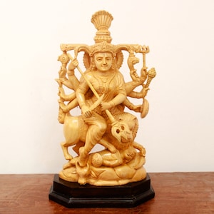 Puede incluir: Una estatua de madera tallada a mano de una diosa hindú con múltiples brazos, montando un león. La estatua está pintada en un color marrón claro y tiene detalles intrincados.