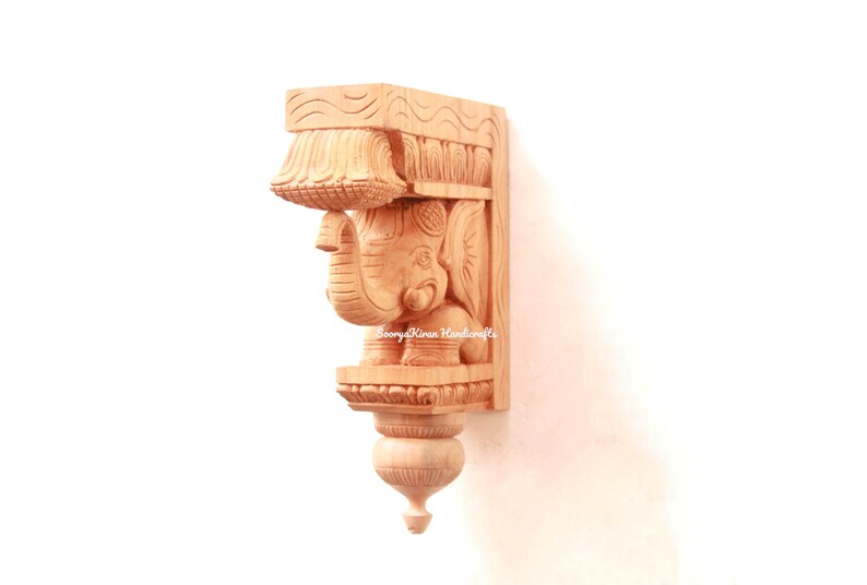 May include: A wooden wall bracket with an intricately carved elephant design. The bracket is made of light brown wood and has a decorative, ornate design.