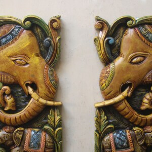 May include: Two intricately carved wooden elephant wall decor pieces. Each elephant is painted in shades of brown, gold, green, and blue with intricate floral designs. The elephants have their trunks raised and are holding a smaller elephant in their trunks.