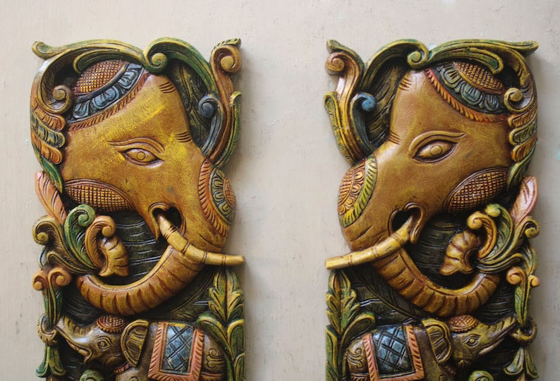 May include: Two intricately carved wooden elephant wall decor pieces. Each elephant is painted in shades of brown, gold, green, and blue with intricate floral designs. The elephants have their trunks raised and are holding a smaller elephant in their trunks.