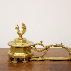 May include: A vintage brass incense burner with a peacock finial. The hexagonal base has decorative etchings and rests on short legs. A curved handle extends from the side. The piece is displayed on a wooden surface.