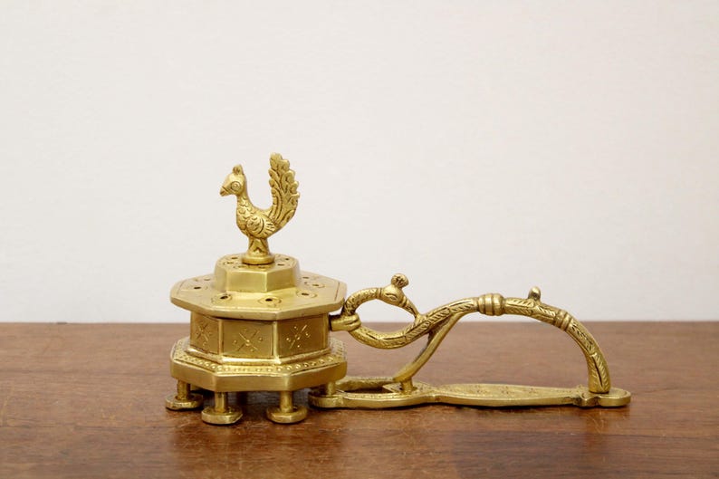 May include: A vintage brass incense burner with a peacock finial. The hexagonal base has decorative etchings and rests on short legs. A curved handle extends from the side. The piece is displayed on a wooden surface.