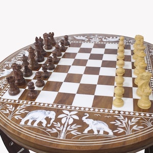 May include: A round wooden chessboard table with intricate carvings of elephants and palm trees. The chessboard is made of dark brown and white squares and features a set of wooden chess pieces in the middle of the board.