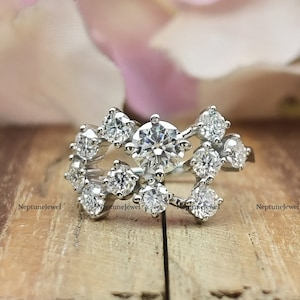 Peut inclure: Une bague en argent avec un groupe de petites pierres rondes et claires. La bague est posée sur une surface en bois avec des pétales de fleurs roses en arrière-plan.