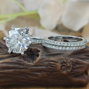 May include: A silver engagement ring set with a large, round, clear center stone and a matching band with smaller, clear stones. The ring is on a piece of brown wood.