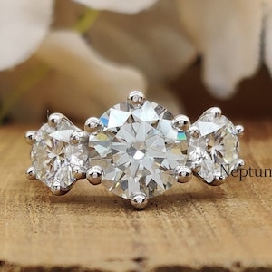 May include: A three-stone engagement ring with a large center diamond and two smaller diamonds on either side. The ring is set in a white metal band.