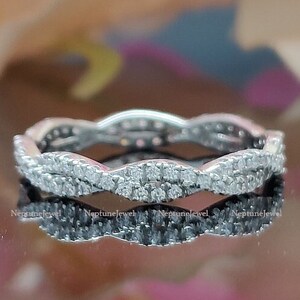 Op de afbeelding: Een zilveren bandring met een golvend ontwerp en kleine, heldere stenen. De ring ligt op een reflecterend oppervlak.