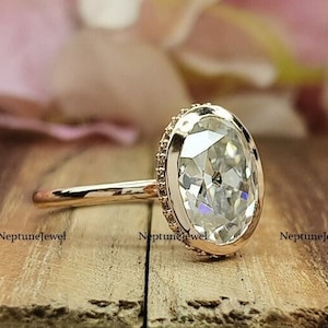 May include: A gold engagement ring with a large oval-shaped diamond set in a bezel setting. The diamond is surrounded by a delicate halo of smaller diamonds.