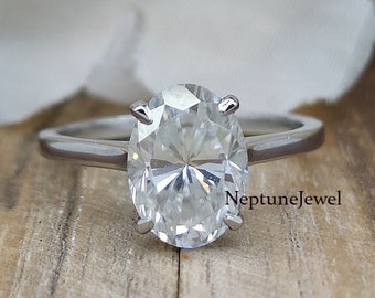 May include: A silver ring with a large, clear oval-shaped diamond. The ring is set on a wooden surface with white flowers in the background.