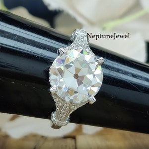 May include: A close-up of a diamond ring with a large, round-cut center stone. The ring has an ornate, detailed setting with smaller accent stones. The band and setting are silver-colored. The text "NeptuneJewel" is visible.