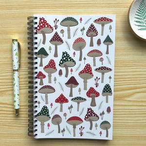 May include: A spiral-bound notebook with a white cover featuring a pattern of red, green, and brown mushrooms. A matching pen with a green leaf design is to the left. A small, round dish with a fern design is in the upper right corner.