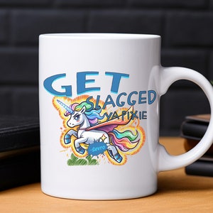 Puede incluir: Taza de cerámica blanca con un unicornio de dibujos animados colorido con una crin y cola de arcoíris. El unicornio está volando con una capa azul y el texto "GET SLAGGED YA PIXIE" en letras azules y amarillas. El texto "Sevro" está escrito en azul debajo del unicornio.