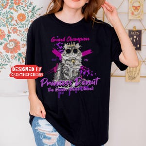 May include: Black t-shirt with a graphic of a cat wearing sunglasses and a crown, with the text "Grand Champion" and "Princess Donut". The shirt also has the text "Designed by Creatingfictions".
