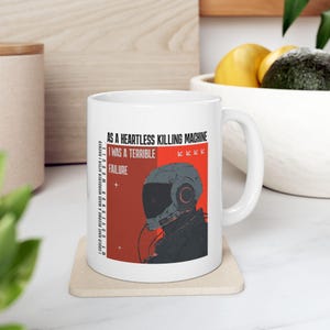 May include: White ceramic mug with a sci-fi themed design. The mug features a gray and red illustration of a helmeted figure and text that reads "AS A HEARTLESS KILLING MACHINE I WAS A TERRIBLE FAILURE". A unique gift idea.