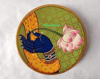 May include: Circular artwork depicting a blue hand holding a pink and white lotus flower. The hand is decorated with gold rings and bracelets. The background is green with a gold geometric pattern and a yellow and gold border.