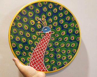 May include: A round wooden plate featuring a hand-painted peacock design. The peacock has a blue head, red body, and green and blue tail feathers. The plate has a gold rim.