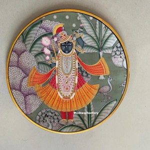 Puede incluir: Una pintura circular sobre un panel de madera que representa a una deidad hindú con un atuendo colorido y adornado de pie en un jardín con pavos reales y otras aves. La pintura está enmarcada en oro.