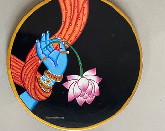 May include: A round wooden plate with a black background and a gold rim. A blue hand with gold bracelets holds a pink lotus flower with a green stem. The plate has an orange and red patterned fabric draped over the top. The text "@pichwalfactory" is at the bottom of the plate.