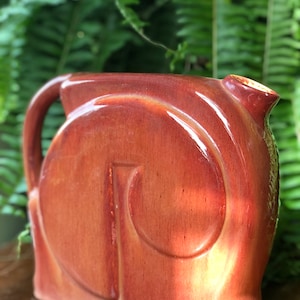 May include: A reddish-brown ceramic pitcher with a curved handle and a spout. The pitcher has a unique design with a large, stylized swirl on the front. The surface has a glossy finish, and the background is blurred green foliage.
