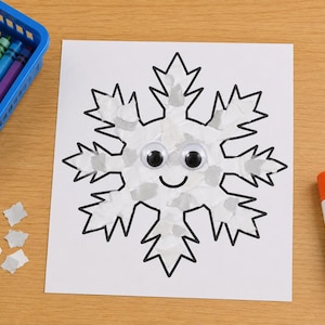 May include: A white paper cutout of a snowflake with black outlines, decorated with small white paper scraps, two plastic googly eyes, and a black drawn smile. A blue plastic basket with colorful crayons and a glue stick are nearby.