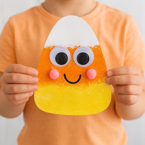 Op de afbeelding: Een handgemaakte candy corn decoratie met wiebelogen, roze pomponwangen en een lachende mond. De candy corn is oranje, geel en wit. Een feestelijke decoratie voor Halloween.